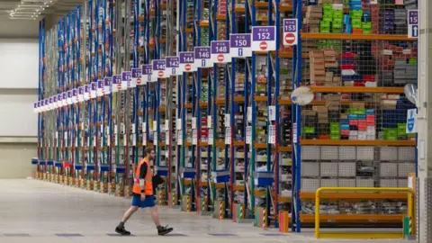 An Amazon warehouse worker walks in front of a vast wall of goods