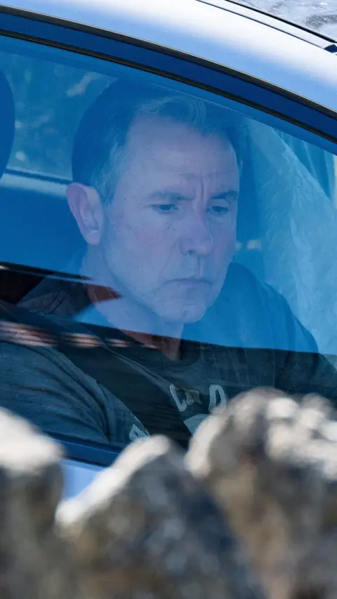 Dan Norris sat in a car, the light is reflecting off the window. In the foreground is the top of a wall.