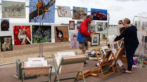 Art on the Prom A man stands in front of a metal fence where he has placed his artwork upon for people to look at and buy. Two women browse through other pieces of his art. 