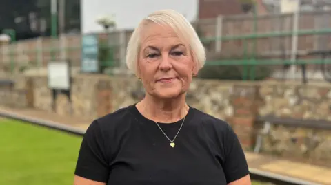 Jack Maclean/BBC A woman standing on a bowling green. She is looking directly at the camera with a serious look on her face. She has short blonde hair and is wearing a black short-sleeved T-shirt with a gold heart pendant necklace round her neck.