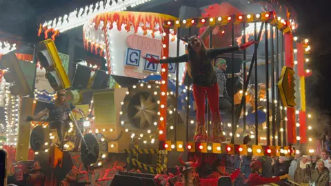 An illuminated carnival cart. There is a woman stood at the front with her arms stretched out.