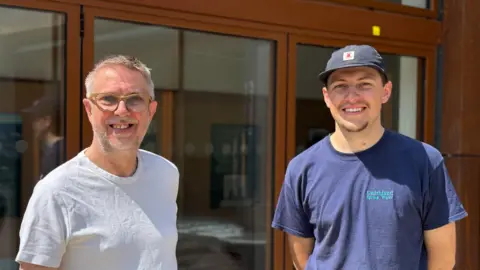 Two men, one wearing a white t-shirt and glasses and one wearing a dark blue t-shirt and cap, standing in front of a glass door.