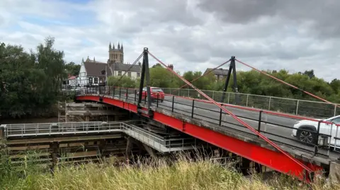 This image shows cars heading into Selby town centre over the toll bridge. The metal sides of the bridge are painted red, as are some supporting cables. The sky above is a mixture of blue and grey. Selby's ancient abbey is in the background.
