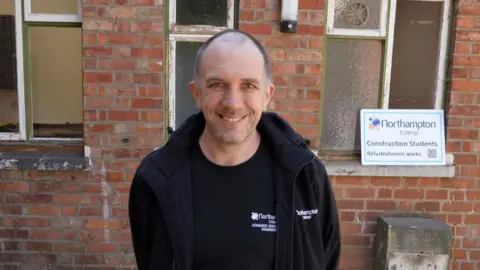 BBC/Qays Najim A man in black T-shirt and fleece with very short dark hair smiles at the camera in front of a brick building and a sign which reads "Northampton College Construction students refurbishment works". 