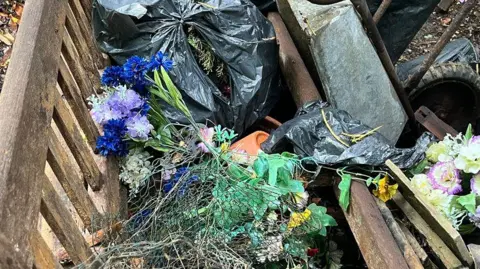 Black plastic bin bags filled with cemetery items sit inside a wooden container, alongside artificial flowers in purple, blue and white, green foliage, broken branches and pieces of mesh and wood, with loose stems and debris scattered among rusted metal and timber.