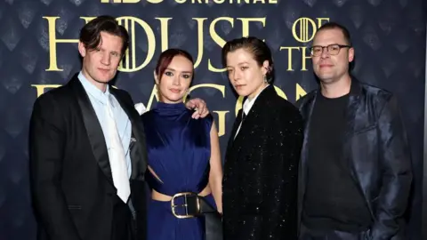 Getty Images Matt Smith, Olivia Cooke, Emma D'Arcy and Ryan Condal stand side-by-side, posing for the camera at a House of the Dragon promotional event. The show's name is written in gold letters on a dark background behind them