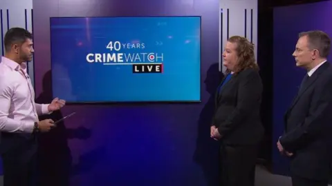 Presenter Rav Wilding to the left of the shot is wearing a pink shirt. There is a TV screen in the middle of the photo with the BBC Crimewatch logo on it, on a blue background. On the right side of the photo are police officers Zoe Hardy and Ian Drummond-Smith, both wearing black suits.