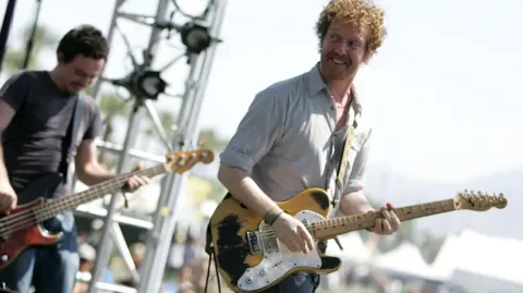 Getty Images Two men on stage playing with a band, the man on the left is holding a yellow and white electric guitar and turning his head back towards a bandmate. He has an amazed smile on his face. Behind him is a man playing a bass guitar, he too is smiling while looking down at the instrument he is playing. It is daylight at a music festival. 