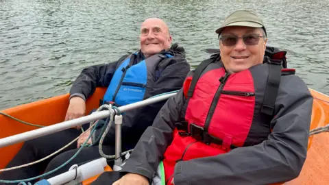 Two men sat in an orange sailing boat on the water. The man on the left has a blue life jacket, dark clothing and no hair. The man on the right has a red life jacket, a dark coat, green cap and sunglasses.