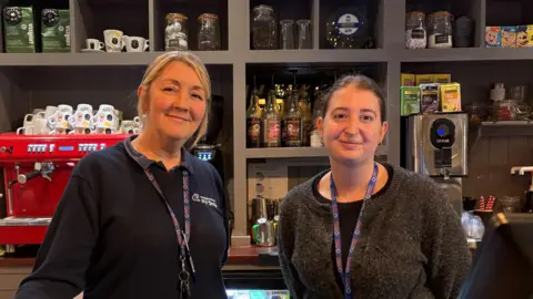Jo (L) has blonde hair, wearing navy blue jumper and around her neck is a work lanyard. Lucy (R) has brown hair, she is wearing a black top with a grey cardigan. Behind the pair is the coffee station where a large bright red coffee machine sits with white mugs on top.