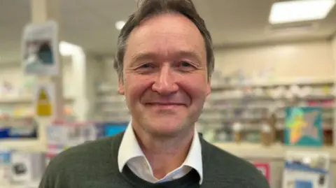 Dr Tim Norbury. He has grey short hair and is wearing a white collared shirt underneath a khaki green jumper. He is pictured looking directly at the camera and smiling. 