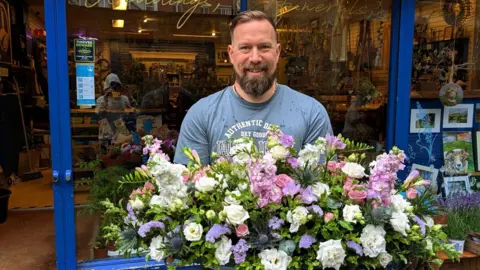 Ant Ratcliffe-Gotts stands outside his shop holding a flower display.