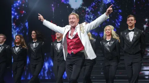Getty Images Michael Flatley holds his arms out wide as he dances on stage. He has short blonde hair and is wearing a white jacket with gold detailing, a white shirt, red waistcoat and black trousers. There are a line of male and female dancers behind him wearing black tuxedos and white shirts. The background is an on screen firework illumination.