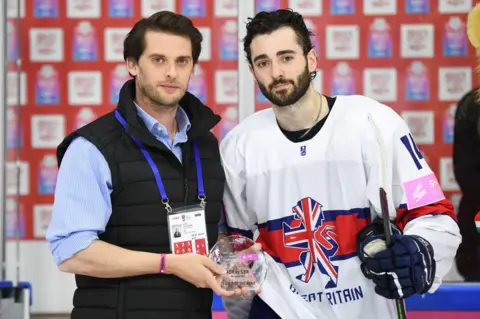 Ice Hockey UK A man in a black body warmer over a blue shirt holds a small, circular glass trophy as he stands next to an ice hockey player in a Great Britain Jersey. The white garment has an outline of a lion - coloured in with a Union Flag design - in the centre.