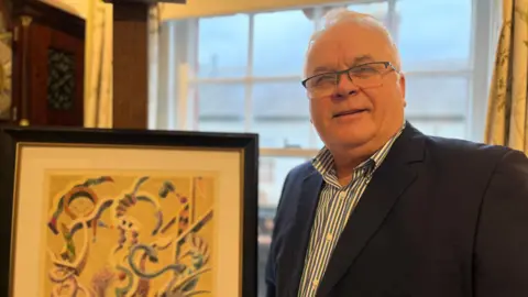 Jan Pietruszka wears glasses, a suit jacket and striped shirt. He looks at the camera while standing in front of a framed Orlik picture.