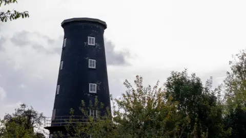 A black windmill with four white windows going down two sides that are in vision. It is situated near several trees and does not have any caps or sails.