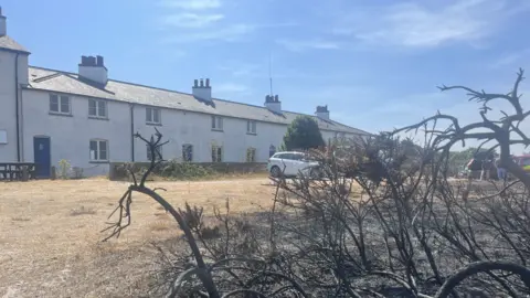 Richard Daniel/BBC A row of white cottages on a sunny day. The ground in front of the cottages has been scorched during a wildfire.
