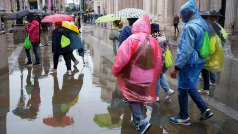 A crowd of people in colourful rain ponchos walk through a rain-soaked street in London. Some are holding umbrellas. 