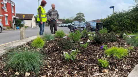 Flowers in a bed with bark. There's green plants peeking through and two people stood inspecting them.