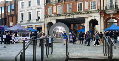 Andrew Woodger/BBC An open paved square in a town centre. A "portal" installation shows people in another country. People on the square are looking at it with one waving. An archway with "Lloyd's Avenue" written above it is behind the portal. There are a few gazebo stalls on the square, and railings in the foreground