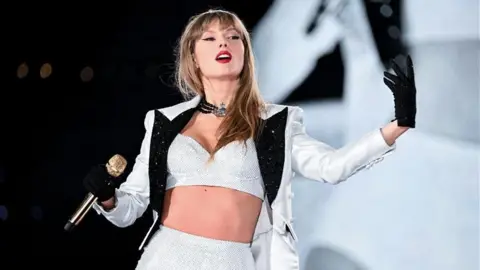 Getty Images Taylor Swift wearing a white jacket and black gloves. She is holding a gold microphone.