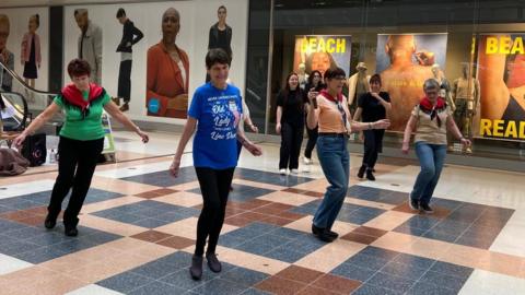 A group of four women perform a line dance routine with shawls on their shoulders in a shopping mall in Crawley 