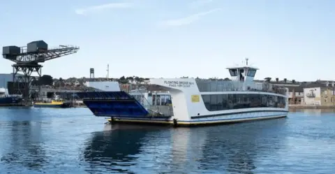 White chain ferry mid crossing on a stretch of river.  To the right on the shore are rows of buildings to the left of the ferry is a large crane.