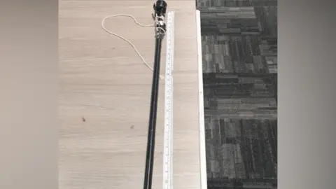 Staffordshire Police A black homemade blowpipe on a table alongside a measuring tape, viewed from above. There is a grey patterned carpet beneath the table.