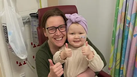 A picture of a woman with a green jumper on and wearing glasses and brown hair smiling into the camera. On her lap is a toddler who has a cream top on and is wearing a purple fabric on her hair. They are both smiling. 