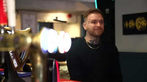 BBC Daniel Williams stands at the bar in a pub. He is a younger man with face and neck tattoos, short brown hair and a beard. He wears a black top and a gold chain.