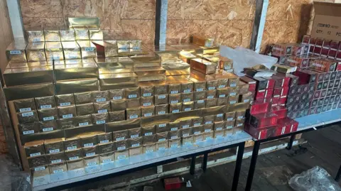 Stacks of gold packages and red packages which look to be perfume packaging stacked on a table.