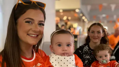 A family sit in a bar enjoying the game. A young woman is wearing her orange Armagh jersey with orange sunglasses on her head. The baby in her lap is also wearing an orange jersey and a spotty bib. Another young woman is wearing a black jersey and the baby on her lap is in orange, sucking on a dummy.