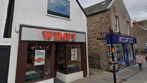 Google The Wimpy is in Dingwall High Street. The front is on the white-painted gable end of a peak roof building. There is a Wimpy sign above the door in red letters.
