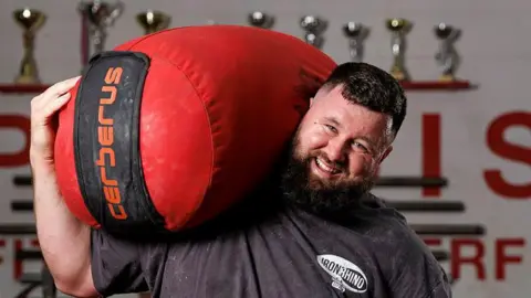 Sgt Anil Gurung Corporal Rhys Calland wears a black T-shirt and a smile as he lifts a huge red sandbag on to his shoulder. 