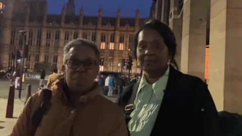 Two women stand in front of Houses of Parliament at night with sad expressions. One wears a brown coat and the other wears a white blouse and black coat.