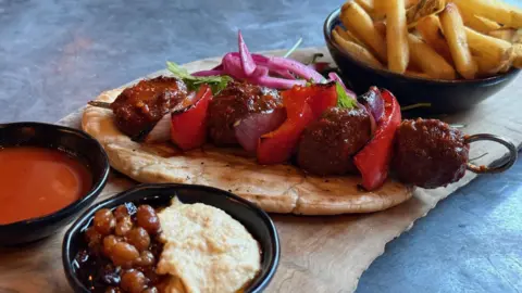 BBC A kebab, with a bowl of chips and a chick pea and houmous dip and bowl of chips alongside.