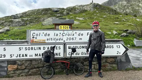 DAVID HOGG a man smiling with a foldable bike in front of signs