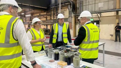 David Macmillan/BBC Labour MP for Redcar and Tees Valley Mayor Ben Houchen, in hard hats, safety glasses and yellow vests talk to scientists in a factory. There are jars and trays of different liquids and materials in a desk.