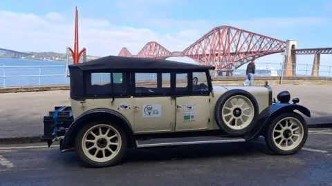 Harry Sear A vintage four-door car parked on the viewing platform by the Forth Bridge near Edinburgh. The red, long-span suspension bridge is in the background over the sea. 