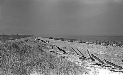 www.erichoskingtrust.com A black and white image showing sea defences along the beach at Minsmere in 1940. On the beach edge in front of the sea on the right is a row of upright and horizontal scaffolding poles forming a grid structure. In front of the sea grass is a row of metal struts pointing out of the sand and with holes in them. 