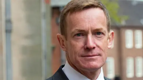 A man with short brown hair looks into the camera. He wears a white shirt and a dark suit jacket.