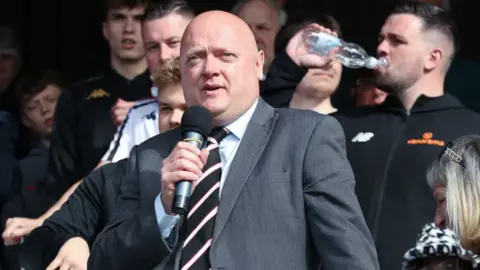 Hereford FC Hereford FC chairman Chris Ammonds , a man in a dark grey suit and balck stripy tie is standing in a crowd holding a microphone while talking 