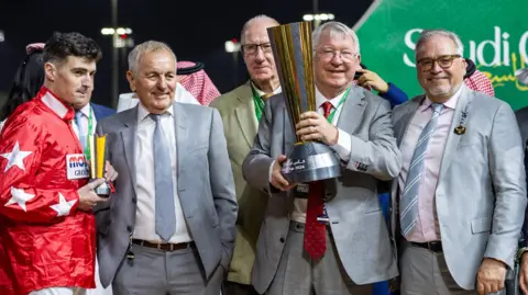Lo Chun Kit/Getty Images Jockey Oisín Orr and owners Peter Done, Sir Alex Ferguson and Ged Mason celebrate with trophies after Spirit Dancer won the Neom Turf Cup in Saudi Arabia in 2024. Orr has short black hair and is wearing a red jockey's shirt with white stars on the sleeves. Sir Alex, who has short grey hair and glasses, is wearing a grey suit and holding a large gold and silver trophy.