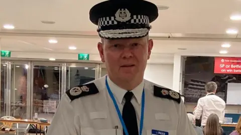 A man in a police shirt featuring decorative epaulettes and wearing a flat police cap.