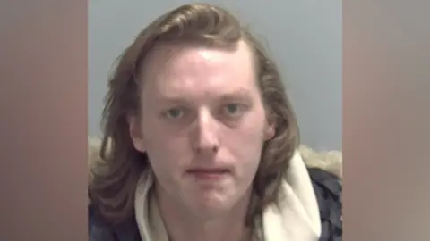 A police mugshot of Owen Trevallion, who has shoulder-length light brown hair and is clean-shaven. He is wearing a grey jacket and a cream fleece.