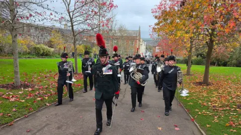 Johnny Rutherford/BBC A parade of band members is seen walking up a path. Trees in autumnal colours line the path.