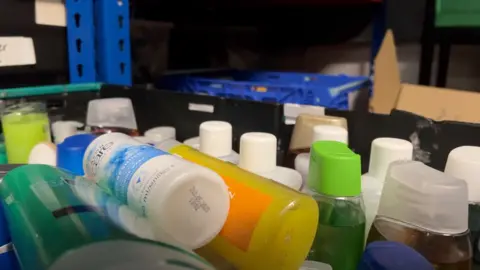 A close-up shot of donated cleaning items in a crate, showing shampoos and shower gels stacked neatly with some extra lying on top. 