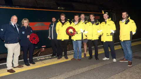 Phil Monckton Six people in yellow coats and three in darker jackets. A woman and a man are each holding a poppy wreath. They are standing on a platform next to the Totnes Castle sleeper train.