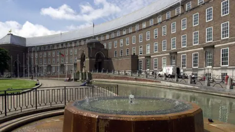 Bristol City Hall at College Green. It is a very large, curved brick building which is brown in colour. There is a fountain at the front and green spaces with people sitting on benches near a path.