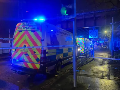 Several police vans under a railway bridge. The area has been taped off following a bus crash